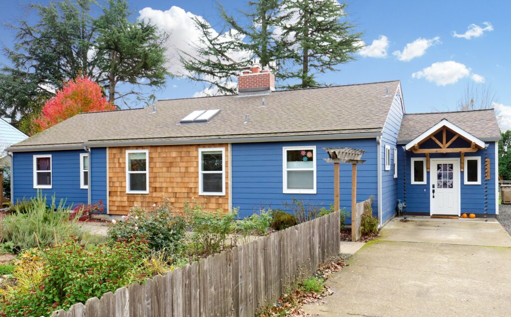 Single family residence exterior with cedar shingle siding and blue paint, wood fence and white entry with trusses, blue sky and trees in background built by Henderer Design + Build Corvallis, Oregon